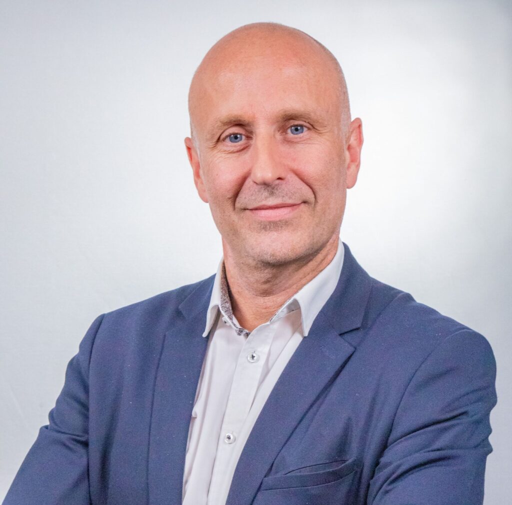 Portrait d'Emmanuel Richard, Directeur du Développement et de la Communication du Groupe ENSIO, veste bleue marine et bras croisés sur fond gris clair