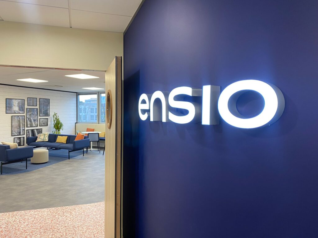 Logo ENSIO rétroéclairé sur mur bleu marine à l'entrée des bureaux, avec espace de convivialité en arrière-plan