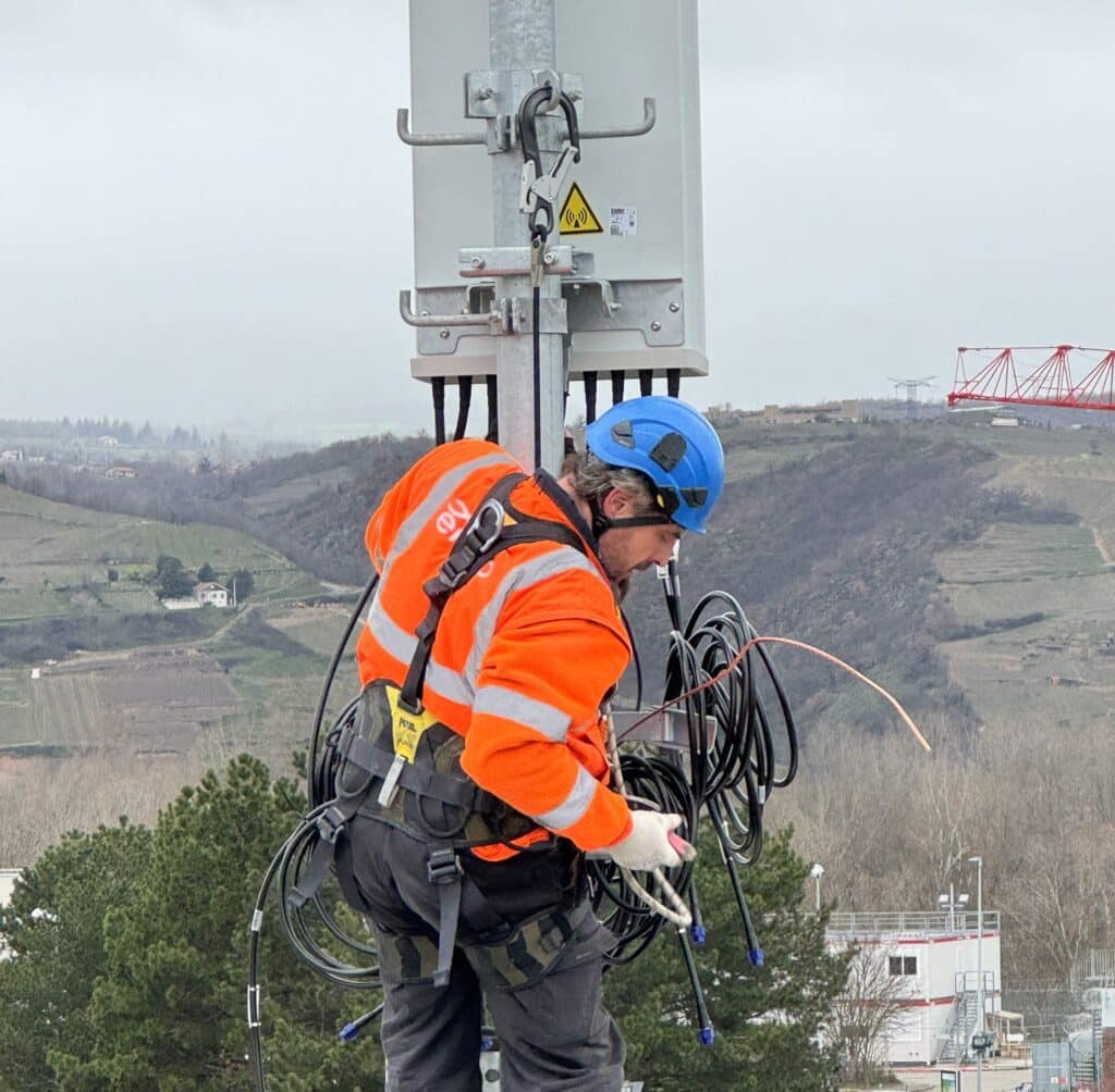 Technicien en veste orange et casque bleu équipé d'un harnais intervenant en hauteur sur un mât télécom avec câbles, sur fond de paysage vallonné