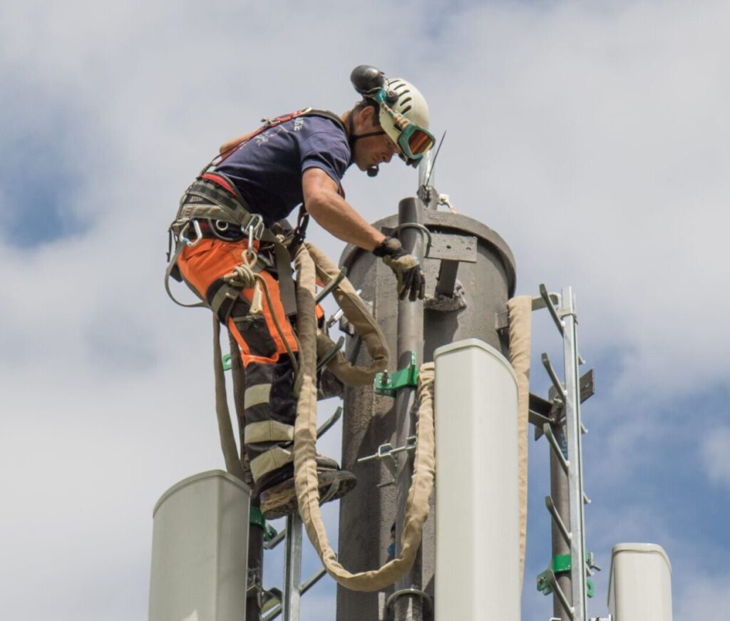 Technicien ENSIO équipé d'un harnais intervenant en hauteur au sommet d'un pylône télécom avec antennes mobiles sur fond de ciel nuageux
