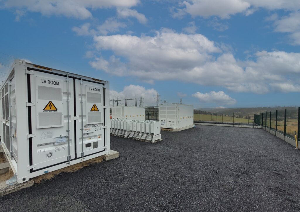 Containers de stockage d'énergie haute tension installés sur un site industriel clôturé avec pylônes électriques en arrière-plan