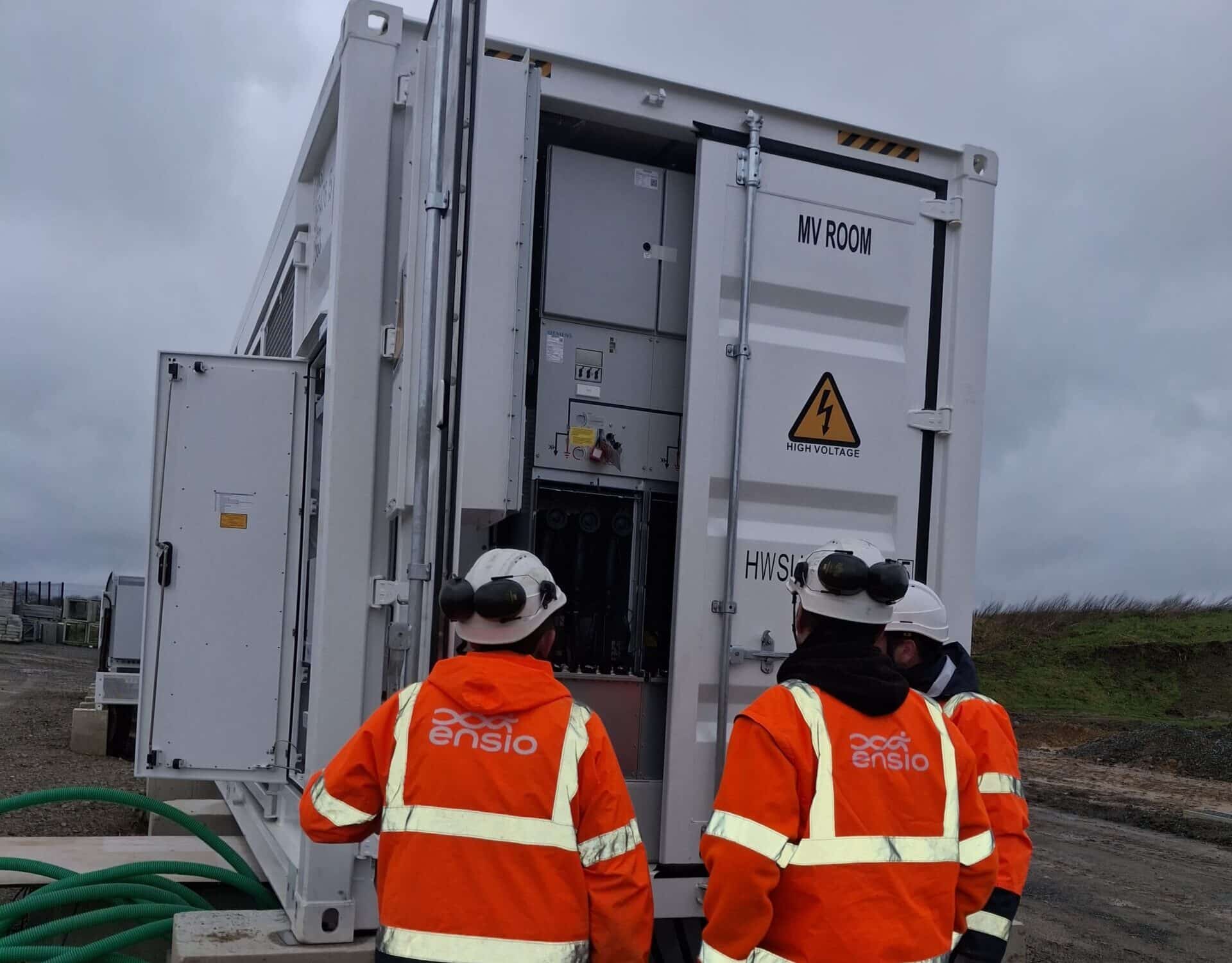Deux techniciens ENSIO de dos en veste orange inspectant l'intérieur d'un container de stockage d'énergie moyenne tension sur un chantier BESS