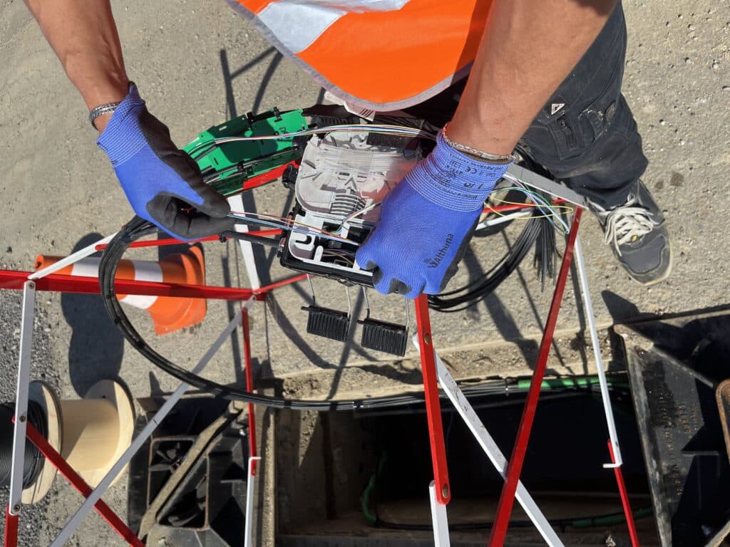 Mains gantées d'un technicien ENSIO manipulant un boîtier de raccordement fibre optique au-dessus d'une chambre de tirage sur un chantier télécom
