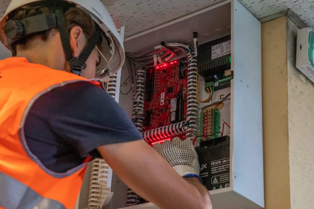 Technicien en veste orange et casque avec visière intervenant sur un coffret électronique mural avec carte électronique et batterie de secours